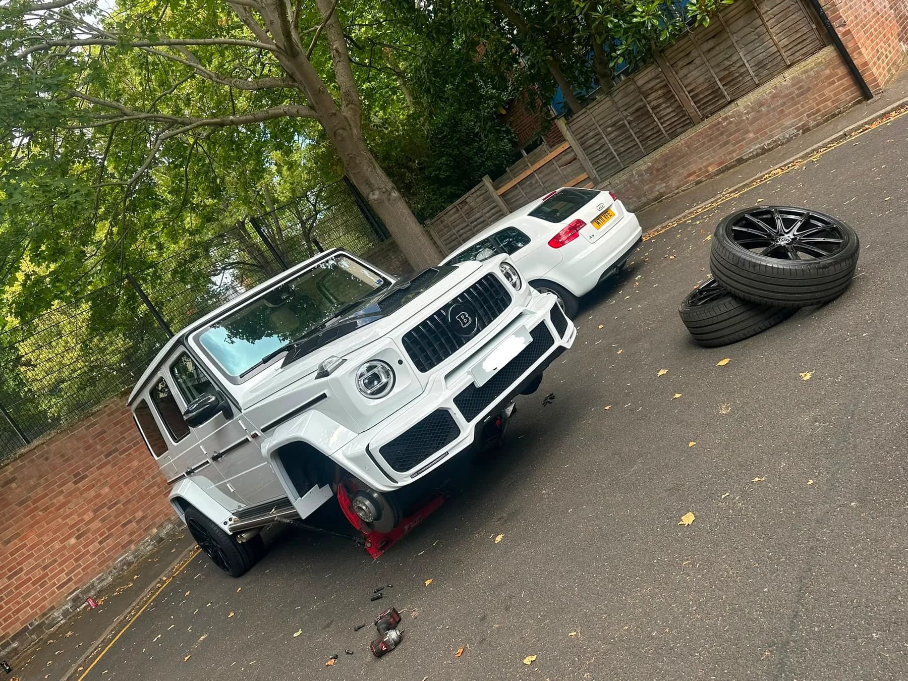 White off-road vehicle raised on a jack in a side street, with wheels stacked nearby